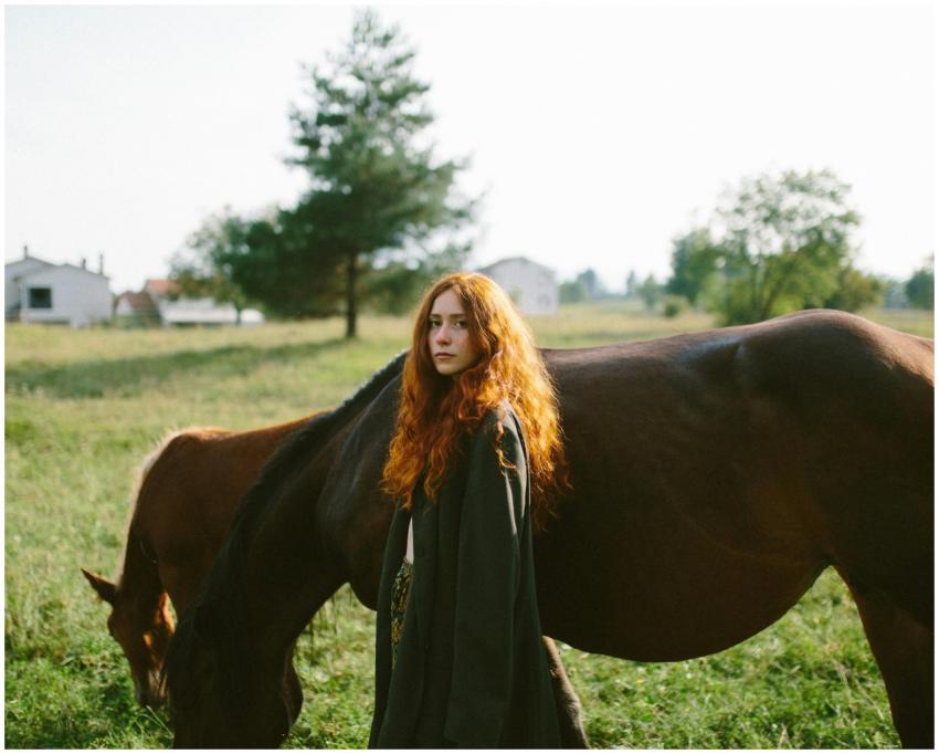 Woman with red hair stands beside a horse in a sun