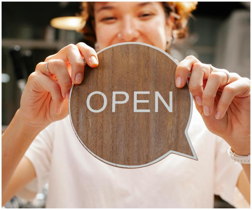 Happy woman holding an 'Open' sign at a café, invi