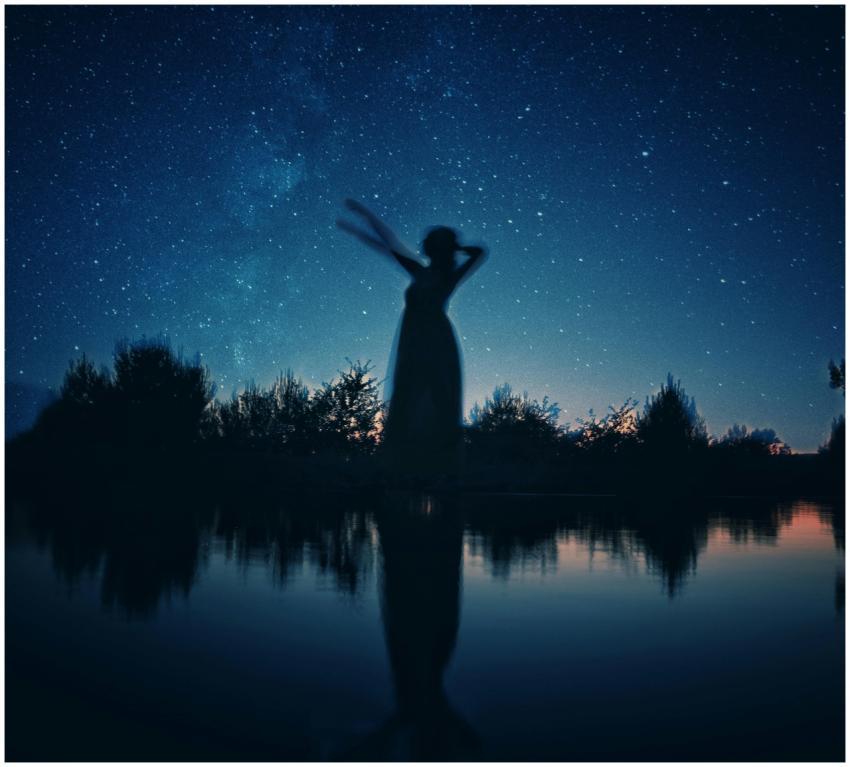 A silhouetted woman stands by a lake under a star-