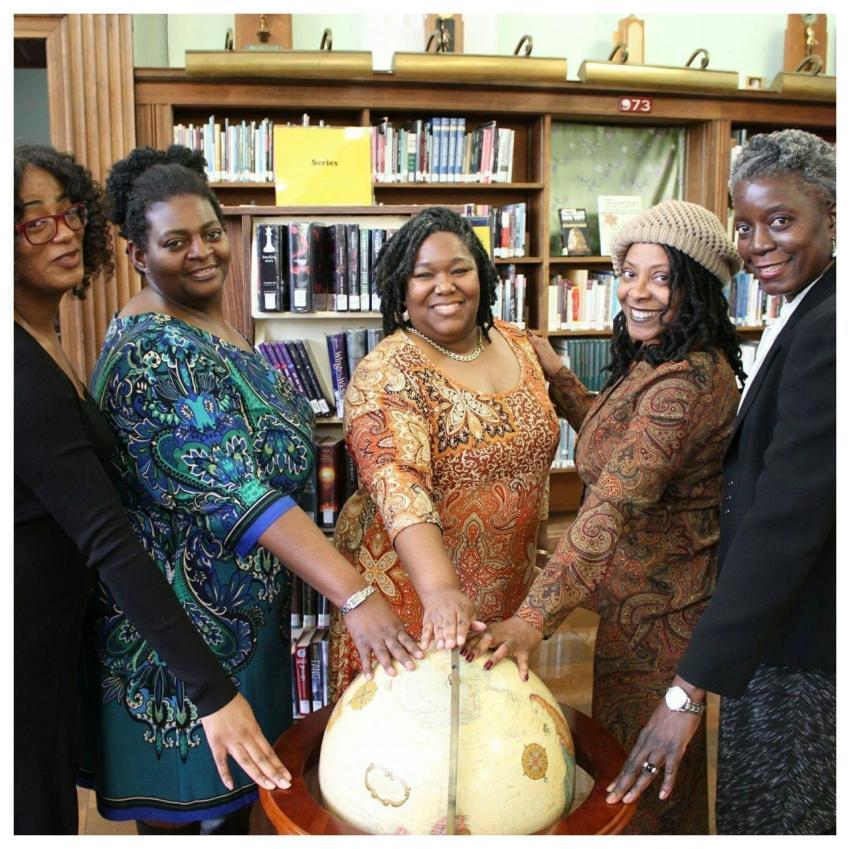 Five women gather around a globe in a library, sym