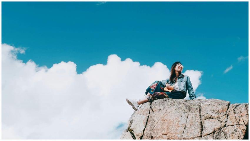 A young woman sitting happily atop a sunny rock un
