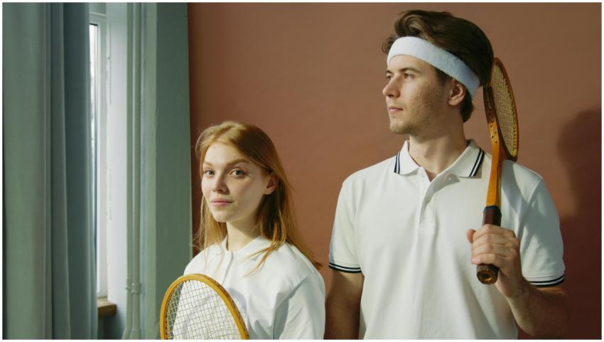 A young man and woman stand indoors with tennis ra