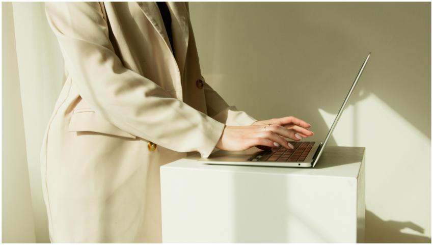 Woman in beige coat typing on laptop in a sunlit w