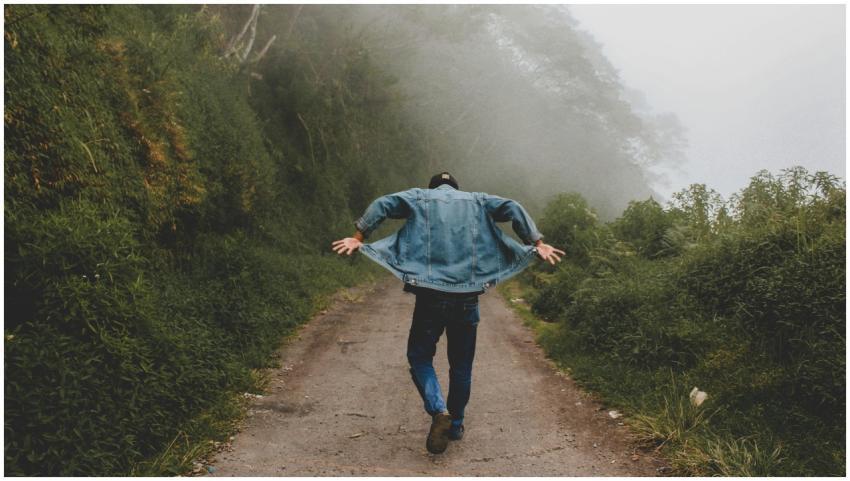 A man in denim jacket hikes along a foggy trail su