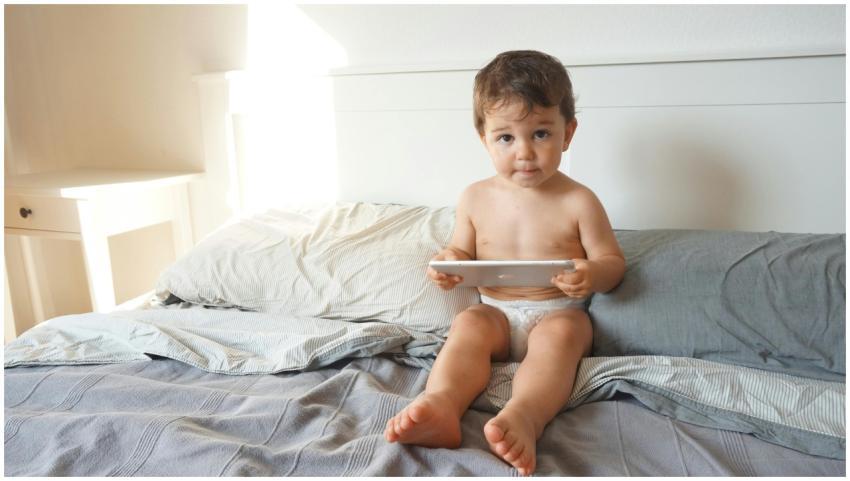 A cute baby sitting on bed with tablet, enjoying a