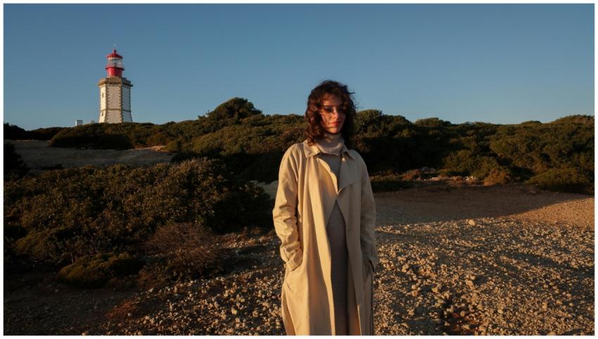 A woman in a trench coat stands near a lighthouse