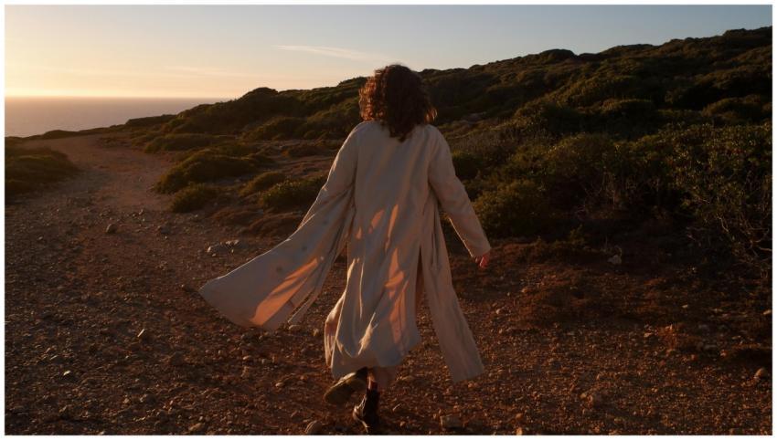 A woman in a coat walks on a scenic coastal dirt r
