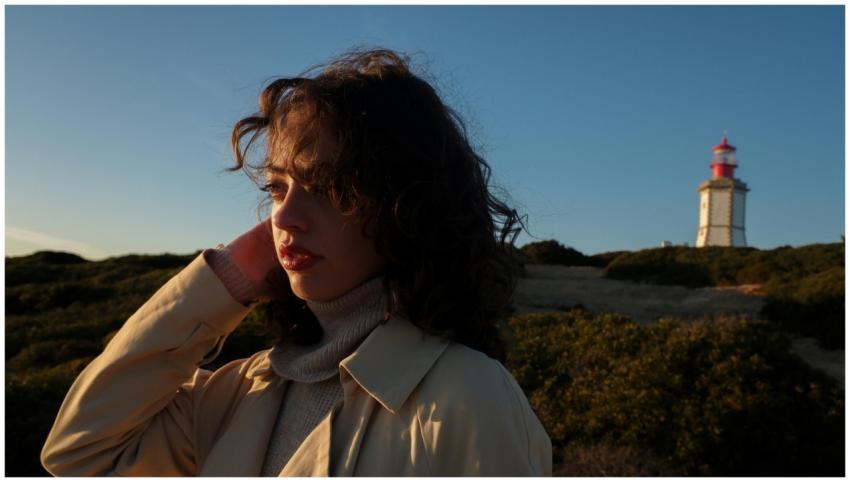 A woman in a beige coat near a lighthouse at sunse
