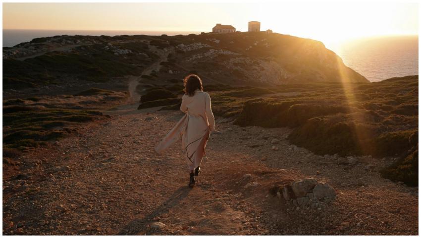 A woman strolls along a rugged cliff at sunset, en
