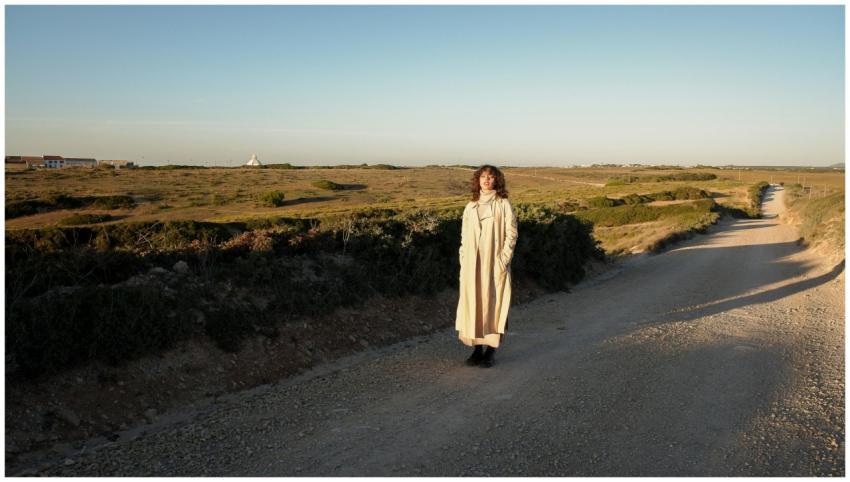 A woman in a beige coat walks on a rural dirt road