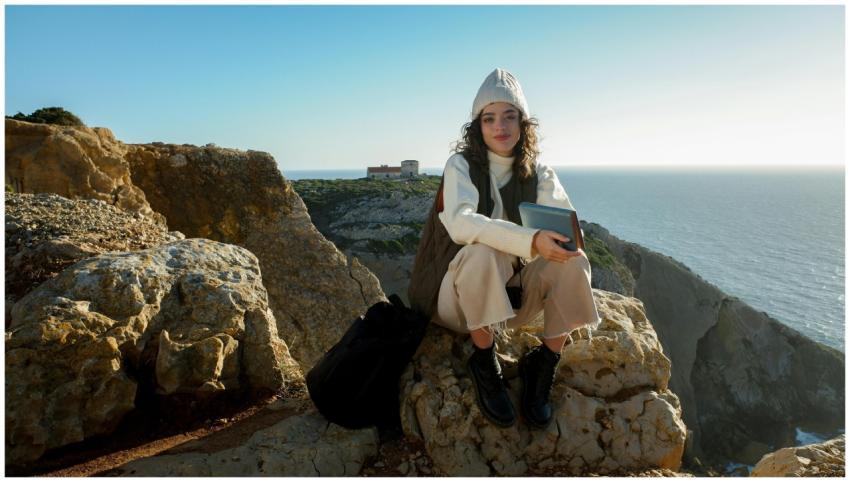 A woman sits on a rocky cliff with a book, enjoyin