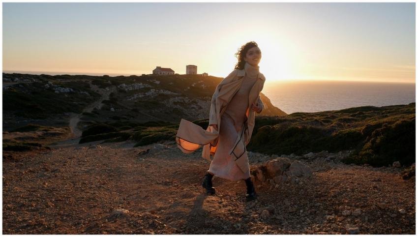 A woman in a beige coat walks by the ocean cliffs