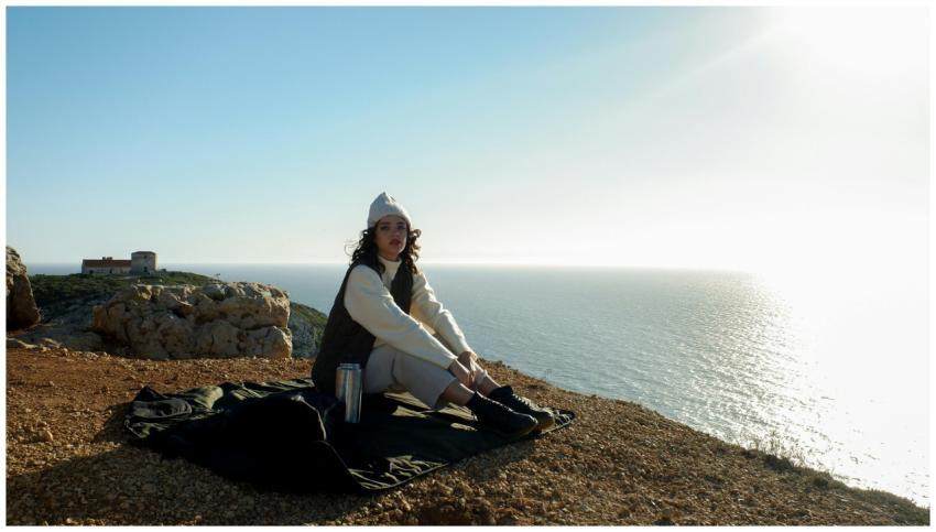A woman in winter clothing sits on a cliff in Lisb