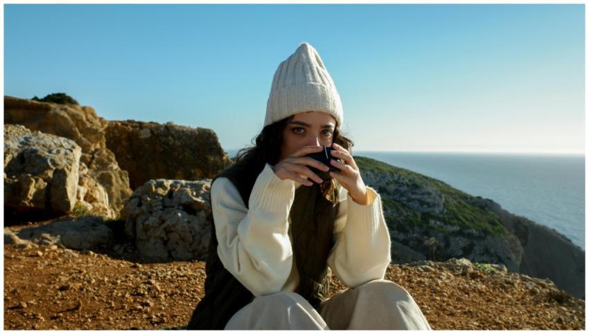 A woman in a cozy beanie drinks coffee by the ocea
