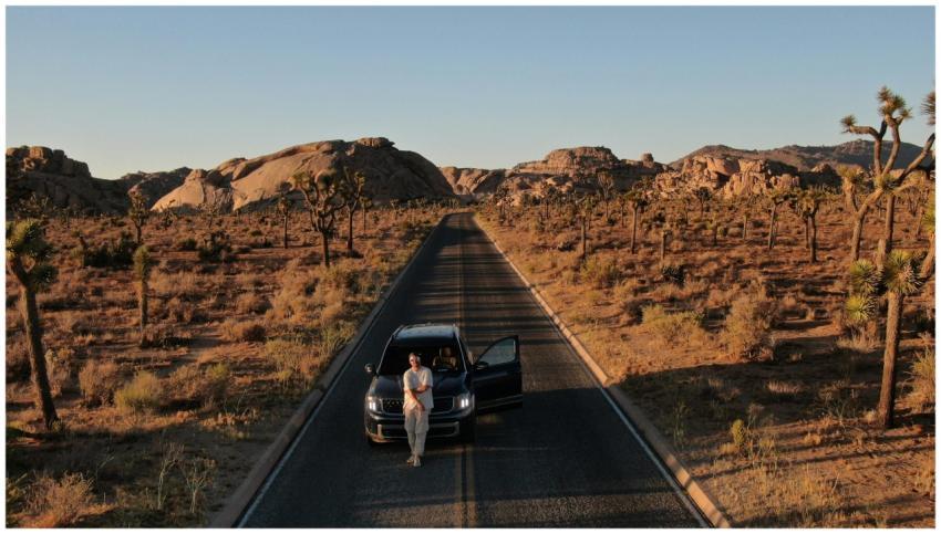 A man leans against a car on a deserted road in Jo