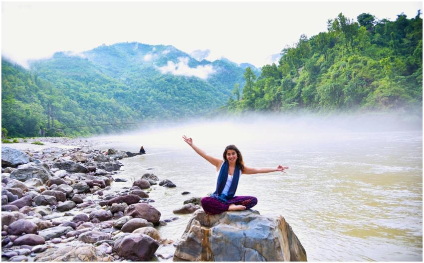 A smiling woman sits on a rock by a misty river, s