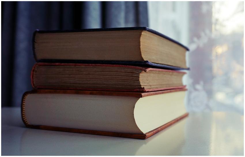 A stack of old books sits on a shelf by a sunlit w
