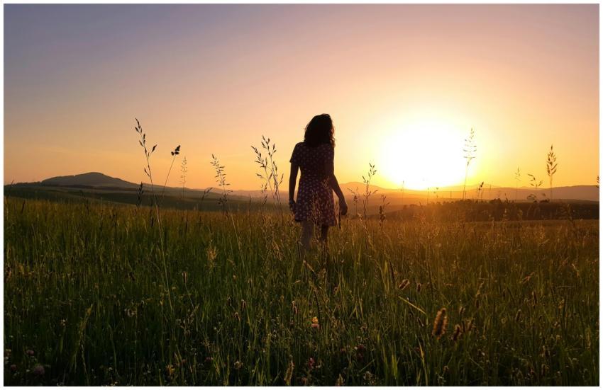 A woman in a field faces a vibrant sunset, capturi