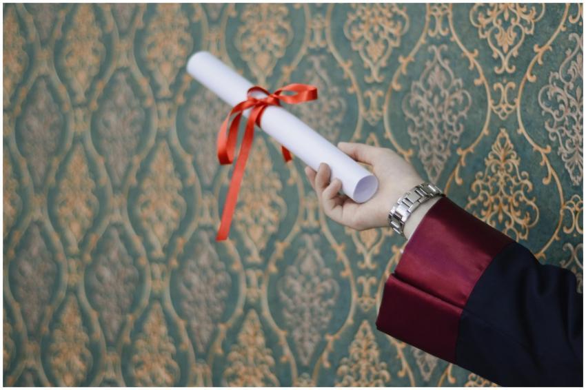 A graduate's hand holding a diploma with red ribbo