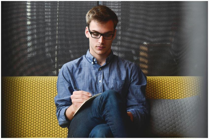 A young man in glasses writes in a notebook while