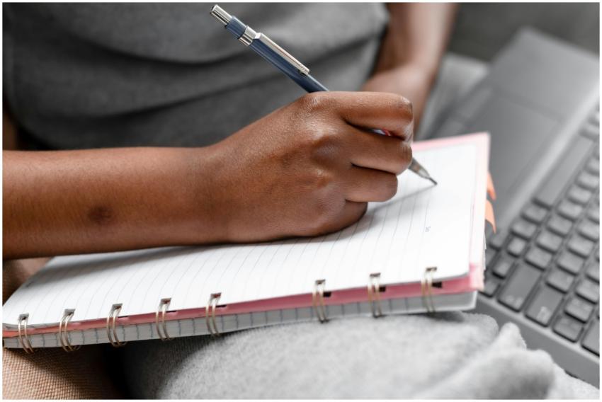 A person writing in a spiral notebook beside a lap