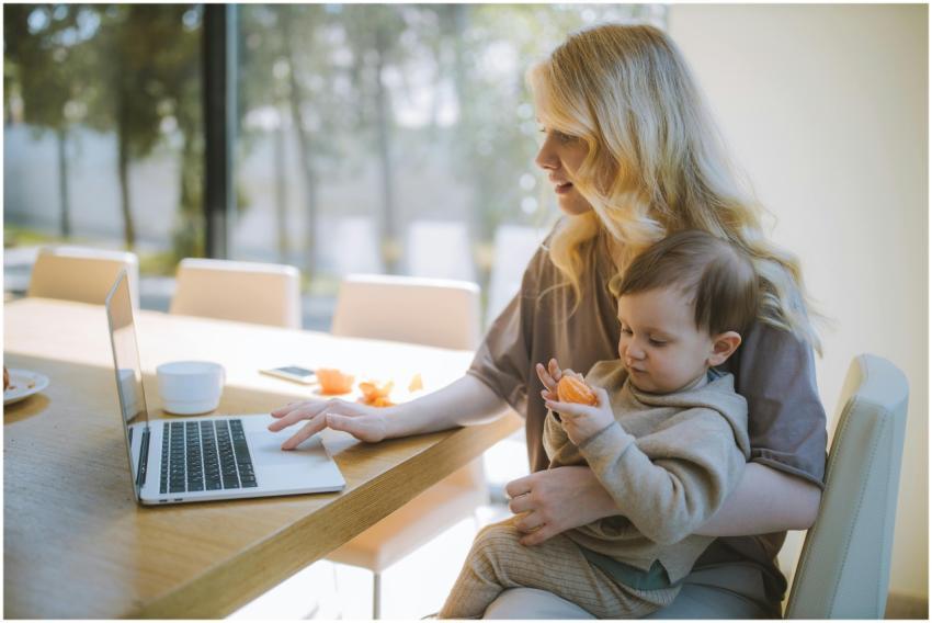 A mother works on a laptop at home while holding h