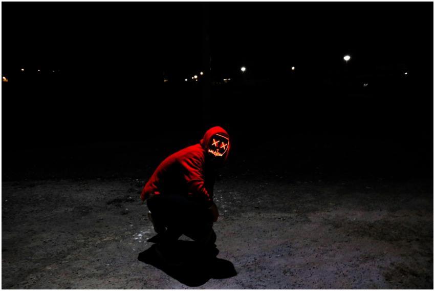 A person in a red hoodie with a glowing mask crouc