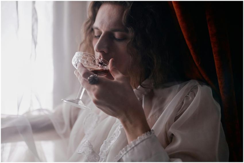 A woman elegantly sips wine in soft light by a win
