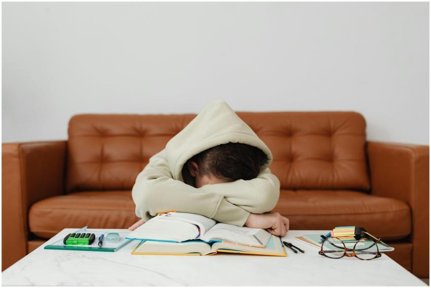 Child in hoodie leans over books, exhausted while