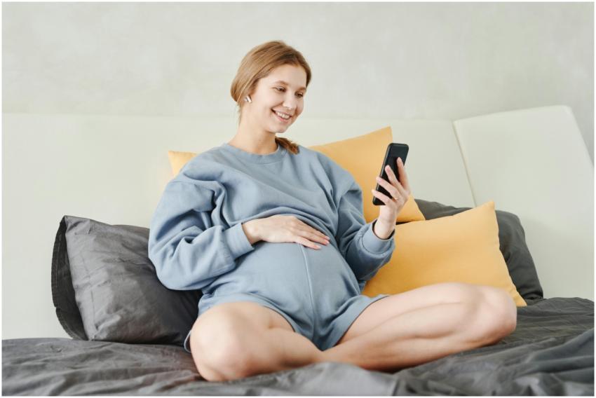A pregnant woman sitting on bed smiling while usin