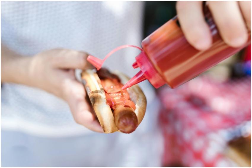 Person adding ketchup to a grilled hot dog held in