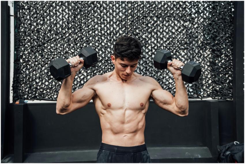 A shirtless muscular man lifts dumbbells at a gym