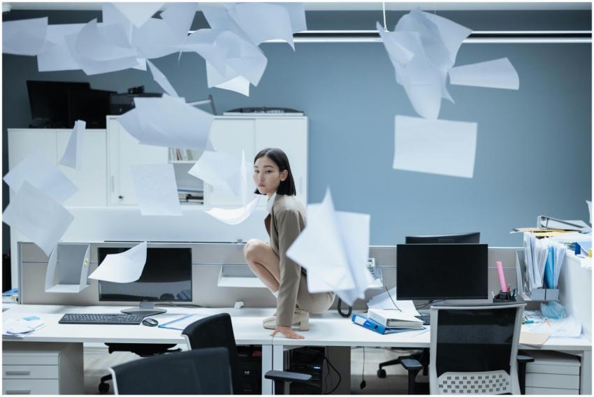 Woman perched on a desk in a chaotic office with p
