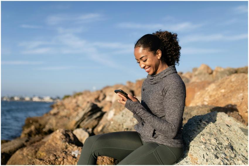 Smiling woman using smartphone on rocky shore, enj