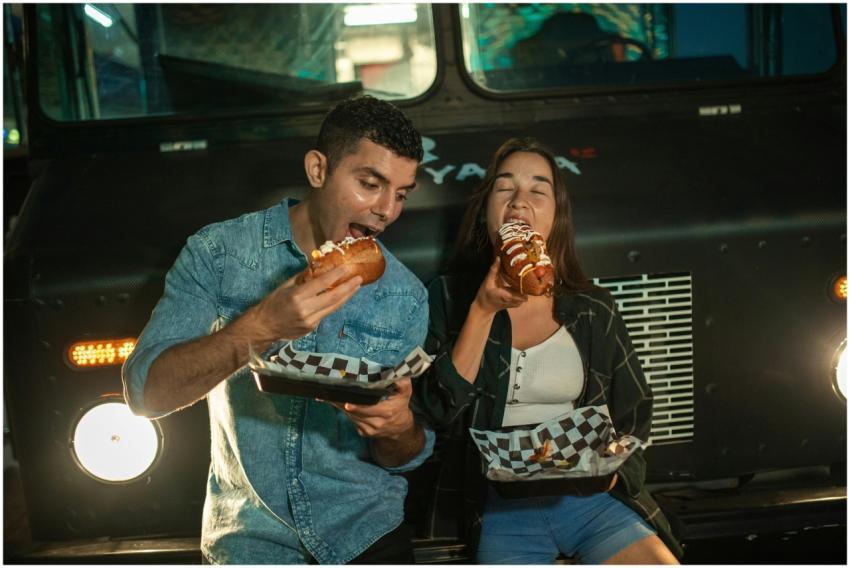 Happy couple eating hot dogs at a food truck, enjo