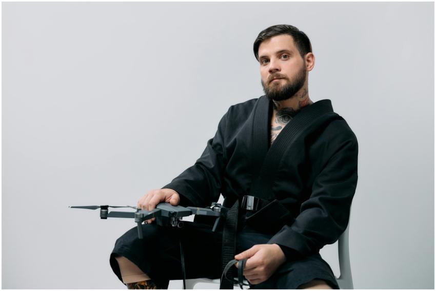 A bearded man sits in a black gi holding a drone,