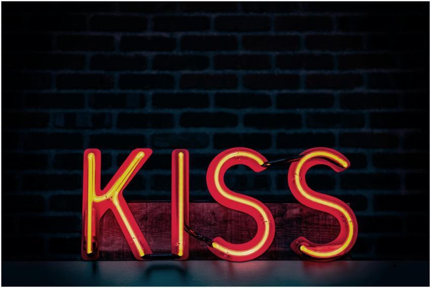 Bright neon lit 'Kiss' sign with a dark brick wall
