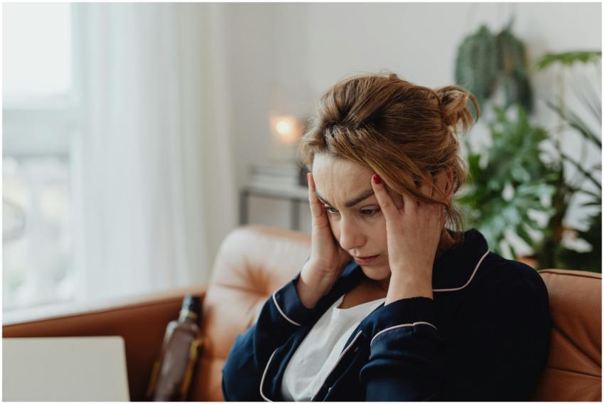 A woman in pajamas sits indoors with hands on head