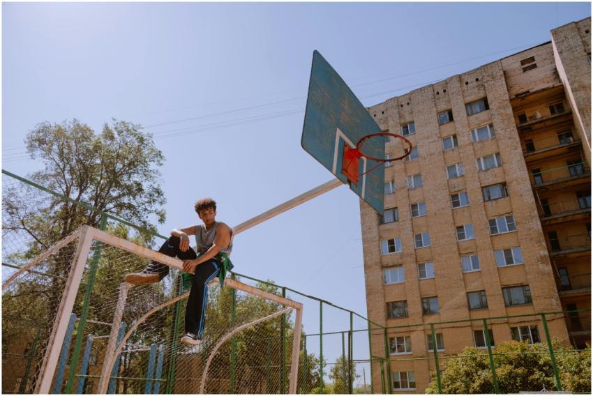 Teenager sitting on a basketball hoop, against a c