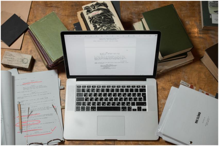 A laptop surrounded by books and scripts on a wood