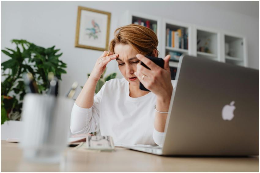 A stressed woman holds her phone, working at a des