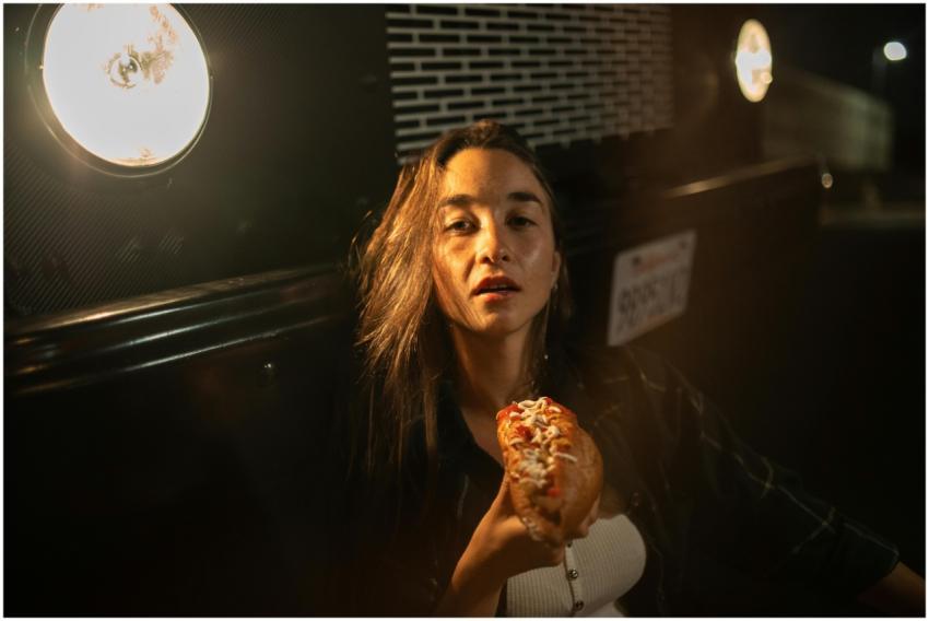A woman enjoys a hot dog beside a lit food truck d