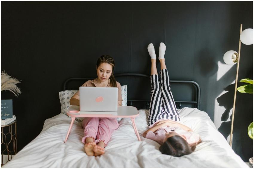 Woman working remotely on laptop in bed, child pla