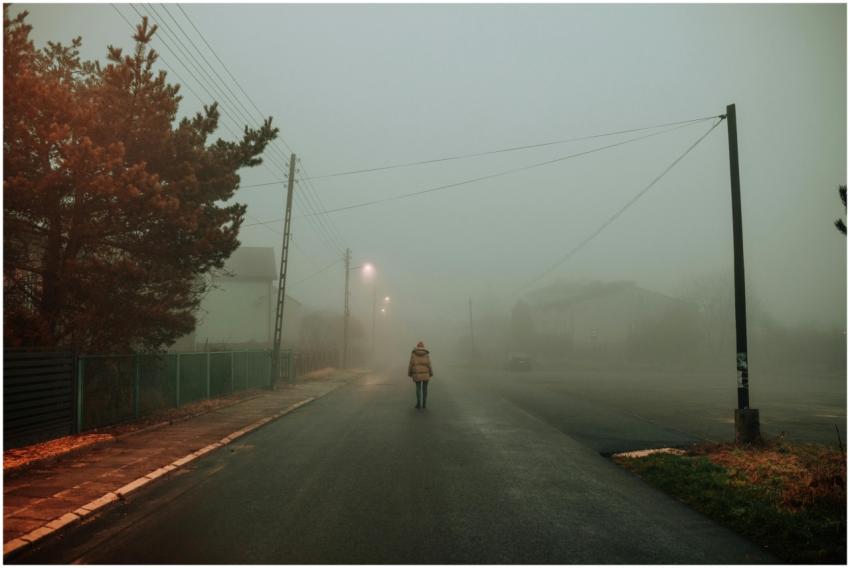 A lone person walks on a foggy, dimly lit street,