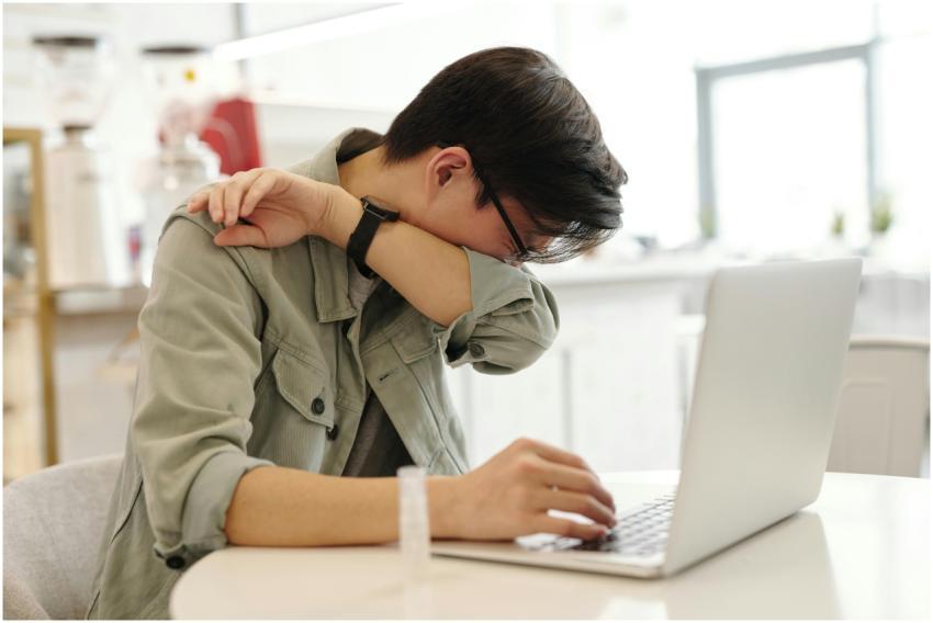 A man sneezes into his elbow while using a laptop,