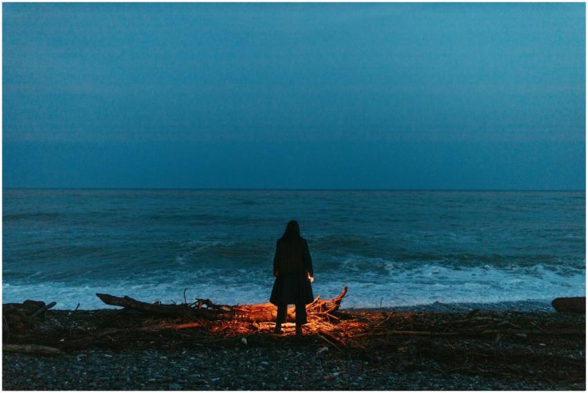 A lone figure stands by a bonfire on a nighttime o