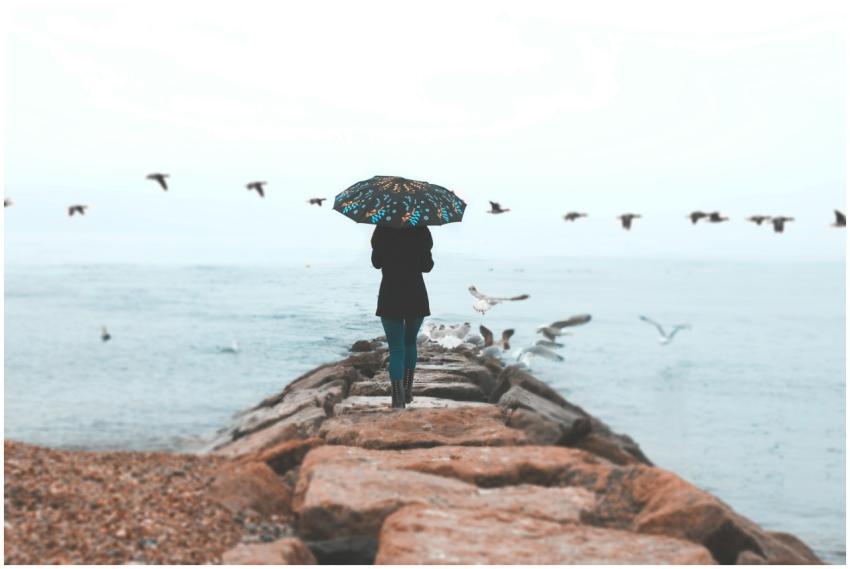 A woman stands on seaside rocks holding an umbrell