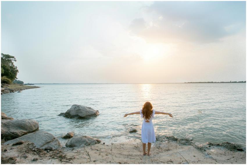 A woman in a white dress stands on a rocky beach,
