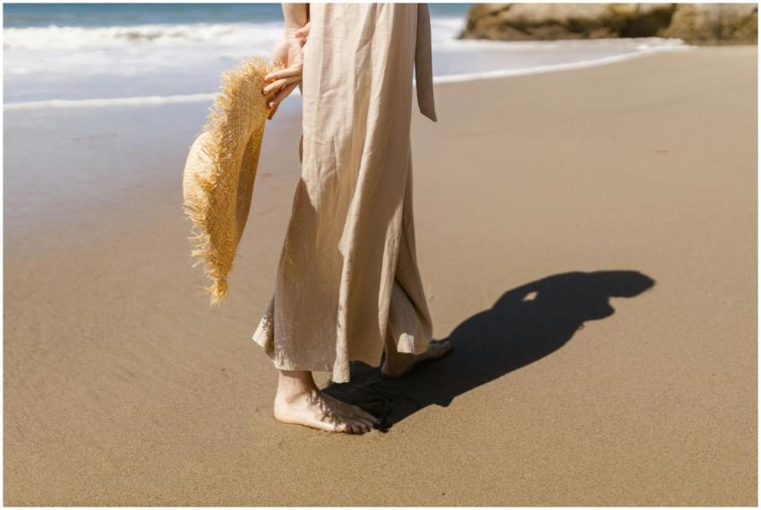 A woman walks barefoot on a sandy beach holding a