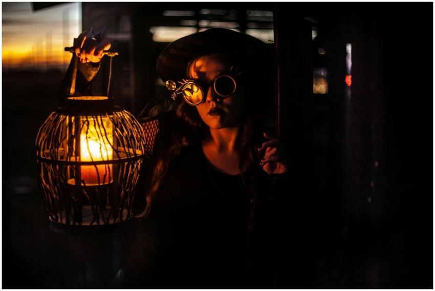 A woman with steampunk goggles holds a lantern, cr